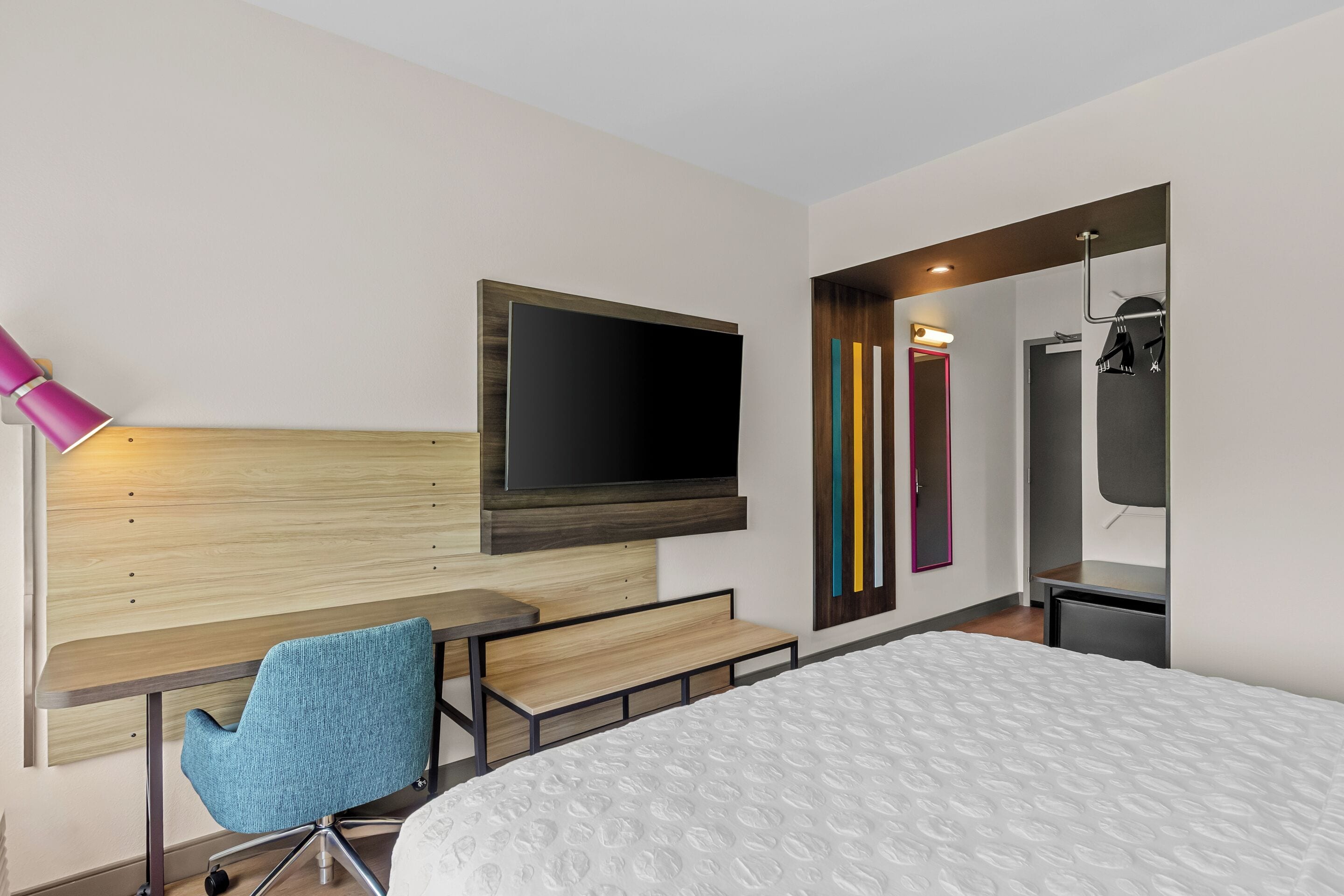 Ein modernes Hotelzimmer mit Bett, Schreibtisch, Stuhl, Wandfernseher und Spiegel.