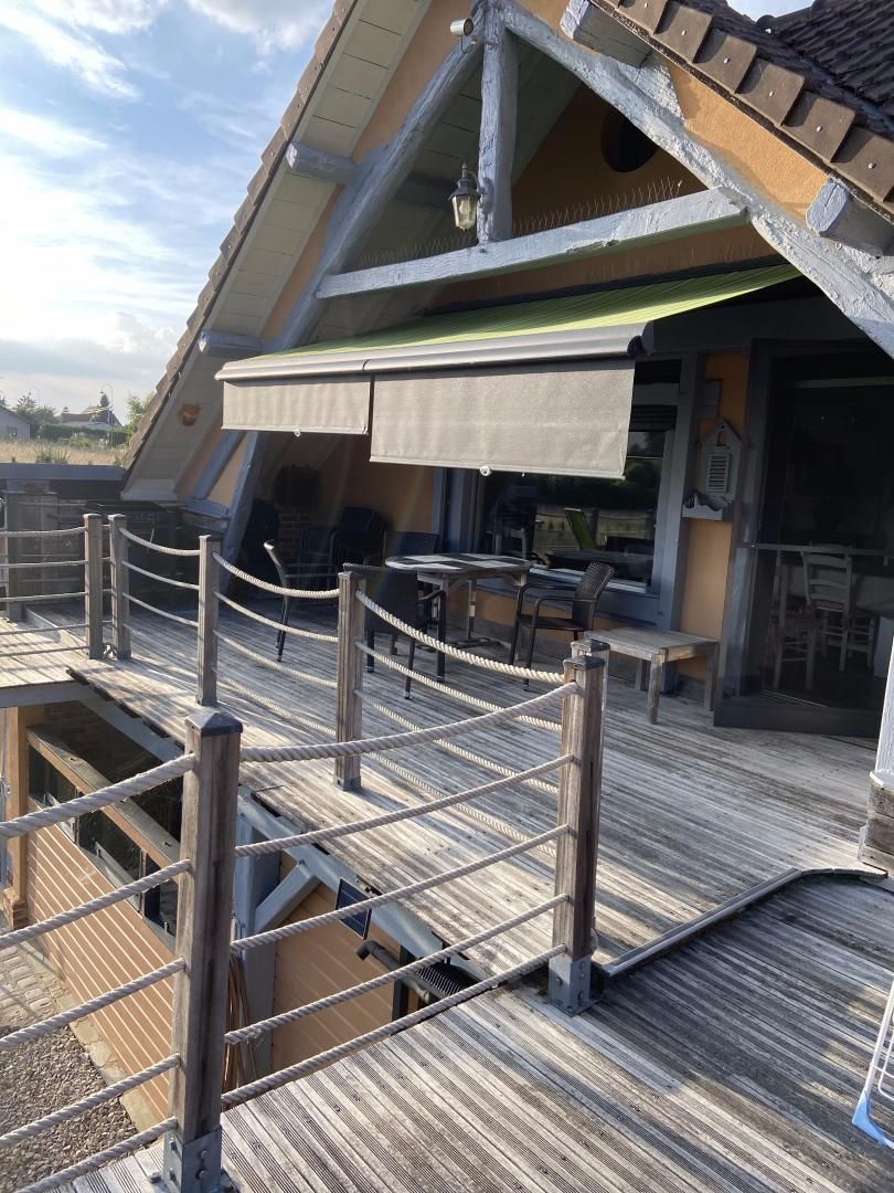 Une terrasse en bois avec une barrière en corde, des sièges extérieurs et un espace abrité surmonté d’une marquise verte.