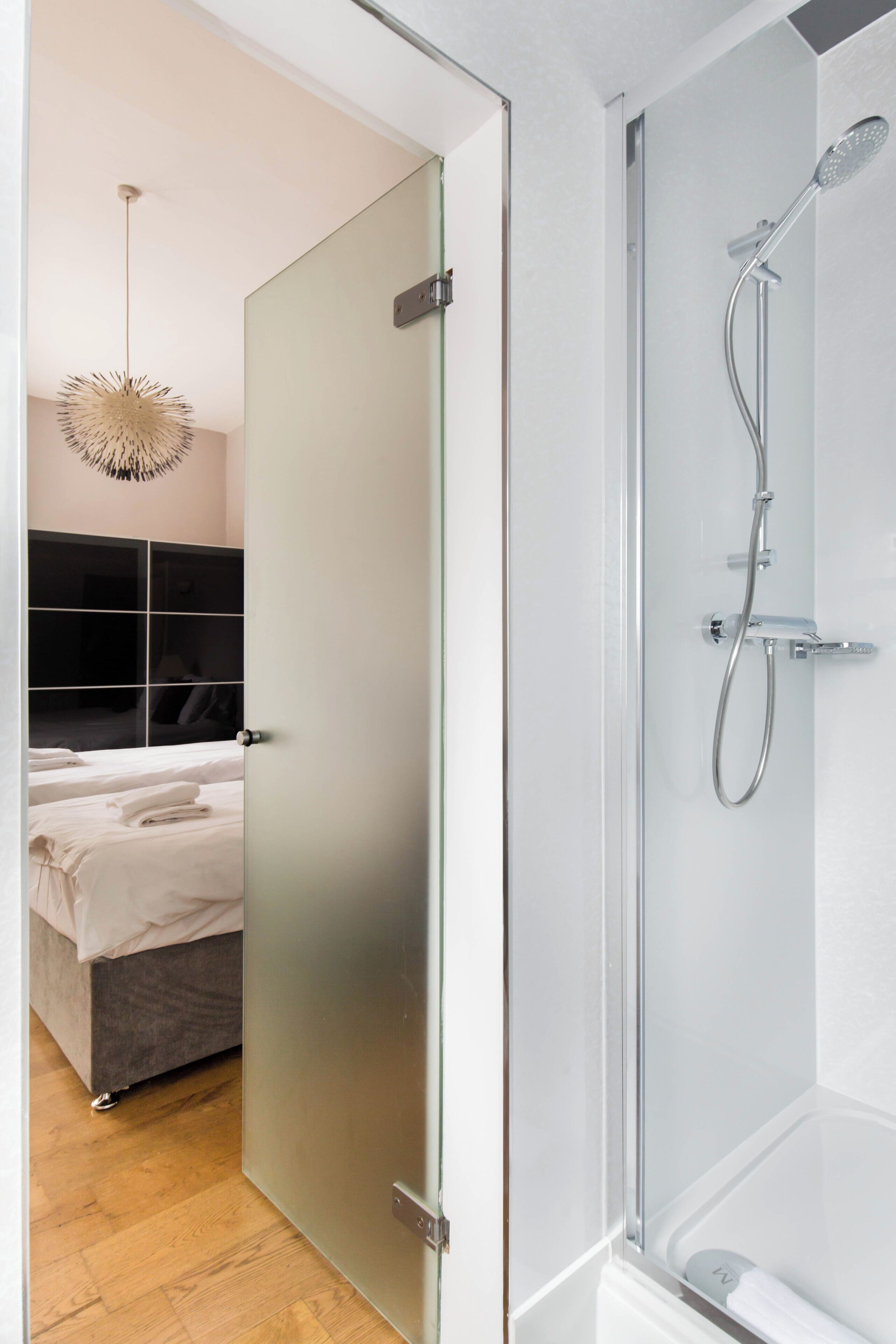 Un baño moderno con cabina de ducha de vidrio, inodoro a la vista y, en la habitación contigua, una cama con ropa de cama blanca.
