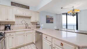 A kitchen with white cabinets, a coffee maker, and a dishwasher. There is a painting on the wall and a view of the sea through a window. - Seaside Beach & Racquet 5914 (Orange Beach)