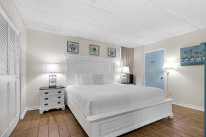 A bedroom with a white bed, two bedside tables with lamps, a door with a blue frame, and a wall with three framed pictures. - Sans Souci Unit 207 (Pensacola Beach)
