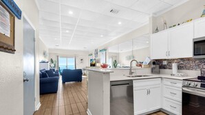 Una cucina moderna con mobili bianchi, una lavastoviglie e un piano cottura. Nell'area living c'è un divano blu e una vista sull'oceano attraverso ampie finestre.