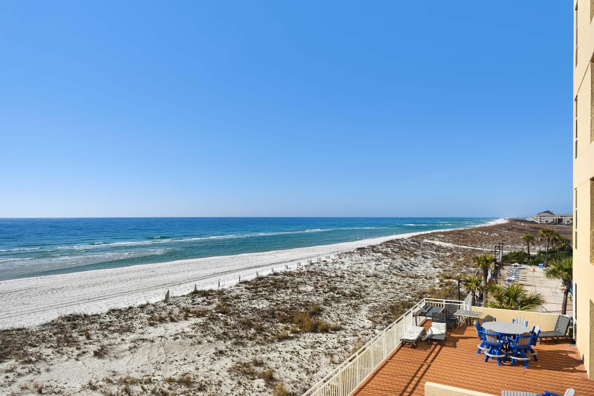 A beach view from a balcony with a wooden deck, outdoor seating, and a clear view of the ocean.