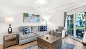 A living room with a grey sofa, a wooden coffee table, a lamp, and a painting of a beach scene. - Sand Dollar E-101 (Pensacola Beach)