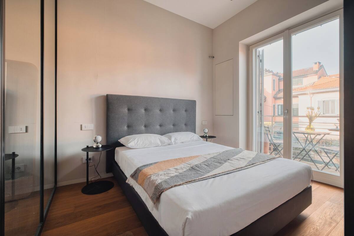 Une chambre à coucher avec un grand lit, une table de chevet, un miroir et une vue sur l’extérieur grâce à une grande fenêtre.