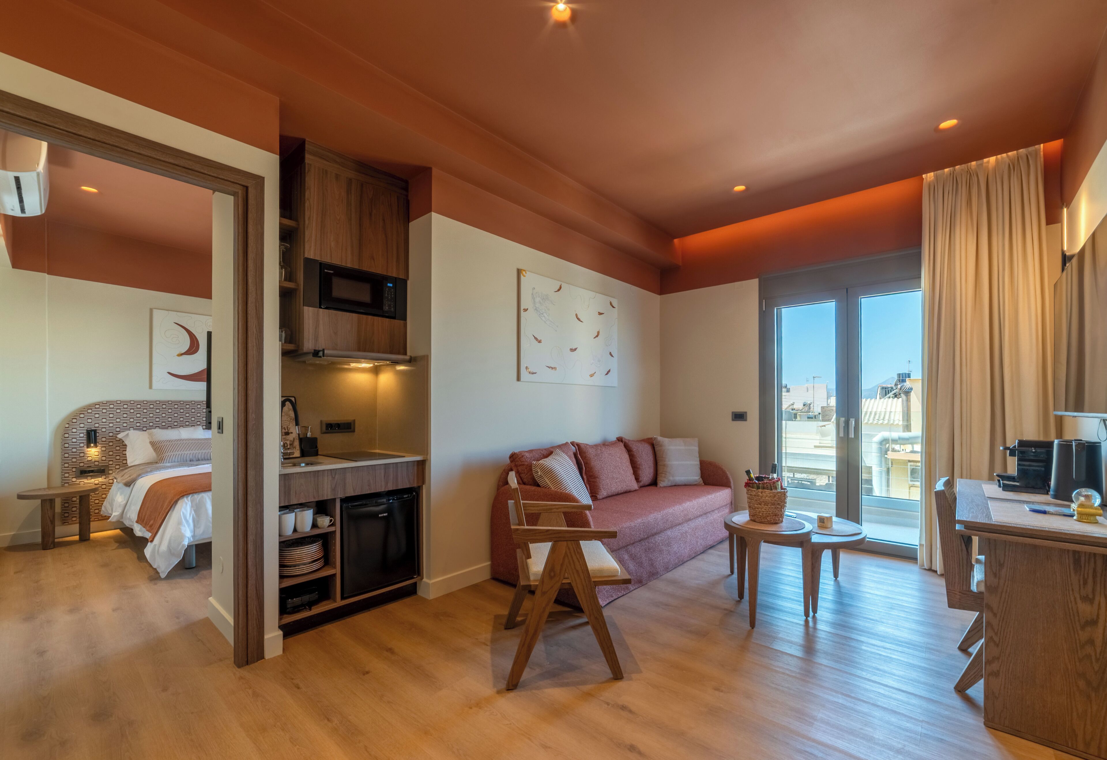 A modern hotel room with a kitchenette, a sofa, a dining area, and a large window.