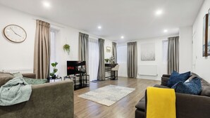 A modern living room with a sofa, a coffee table, and a TV. There is a dining area with a table and chairs. The room has wooden flooring and large windows with curtains. - Comfortable 3-Bed close to Excel Ideal for Groups (London)