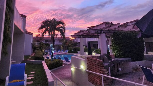 Área de piscina em resort com espreguiçadeiras e uma palmeira, cercada por edifícios e um céu ao pôr do sol.
