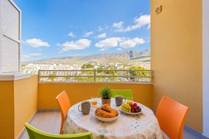 Ein Balkon mit einem für das Frühstück gedeckten Tisch, darunter Croissants, Obst und Kaffee, mit Blick auf eine gebirgige Landschaft.