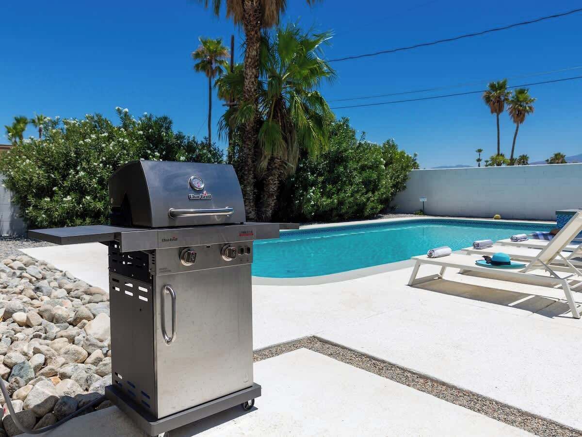 A barbecue grill near a swimming pool with lounge chairs and palm trees in the background.