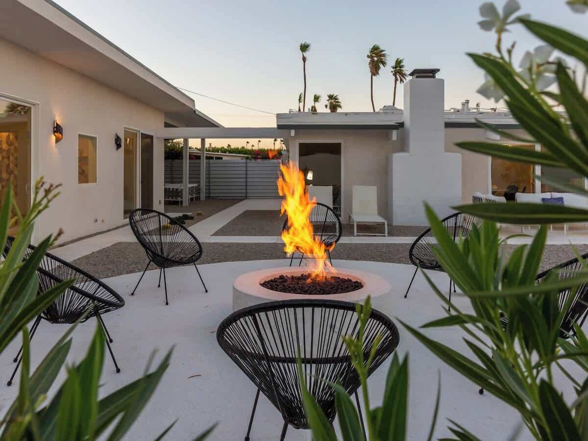 A modern outdoor seating area with a central fire feature, surrounded by black wire chairs, against a backdrop of a white building and palm trees.