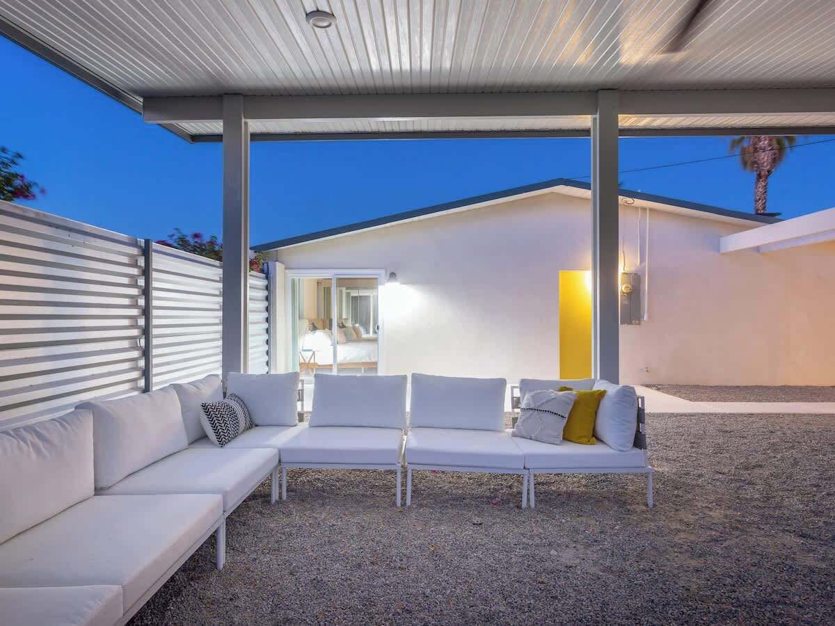 Un'area relax all'aperto, coperta, con un divano bianco e cuscini, pavimento in ghiaia e un edificio sullo sfondo.