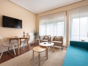Una sala de estar moderna con un sofá, una mesa de centro, una mesa de comedor con sillas, un televisor montado en la pared y ventanas grandes con cortinas traslúcidas.