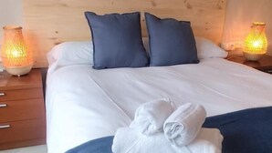 A neatly made bed with white linens and two dark blue pillows, a folded white towel on the bed, and two lit lamps on wooden nightstands.