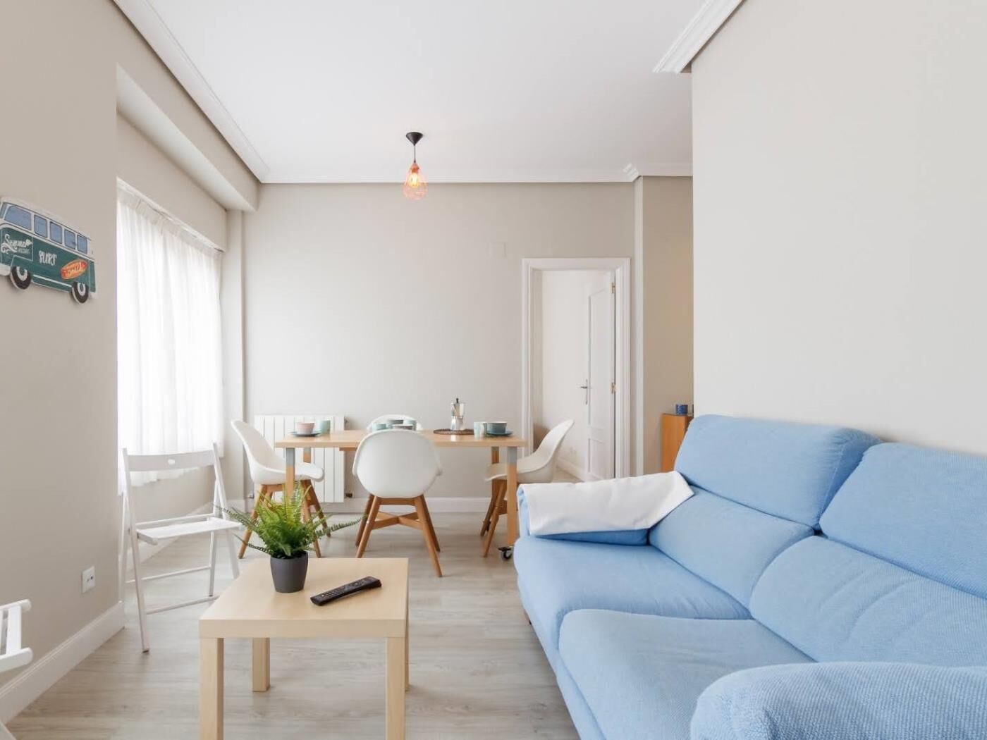 Una sala de estar moderna con un sofá azul, una mesa de comedor con sillas, una mesita con una planta y un control remoto, y un candelabro en el techo.