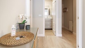 A hallway leading to a bathroom with a sink and cabinets. There is a woven mat on the floor, a potted plant, and a bottle on a glass table.