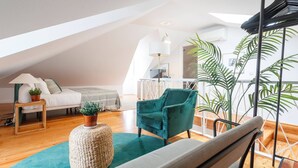 Ein modernes Loft-Apartment mit schrägverdachter Decke, einer gemütlichen Sitzecke mit einem türkisfarbenen Sessel und einer Couch, einem kleinen Holztisch mit Lampe und einem großen Fenster mit Blick auf die Stadt.