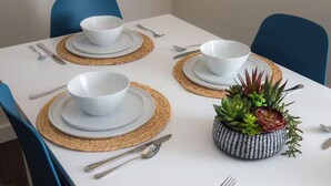 Een eettafel gedekt met witte borden, kommen en bestek, met geweven tafelonderleggers en blauwe stoelen.
