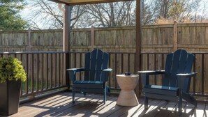 Een overdekt terras buiten met twee Adirondack-stoelen en een kleine tafel, omringd door een houten schutting en bomen.