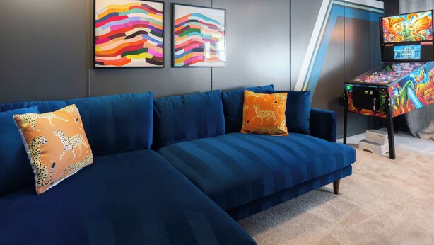 Een moderne woonkamer met een blauwe bank, twee kleurrijke abstracte schilderijen, een speeltafel en een televisie.