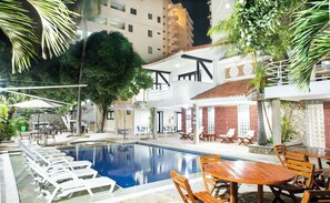 Un espace aménagé au bord de la piscine, avec des chaises longues, une table en bois et un bâtiment doté d’un balcon.