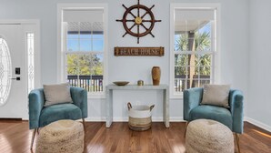Una stanza con due poltrone color petrolio, un tavolino consolle in legno, una ruota di timone e un cartello con la scritta 'Beach House'.