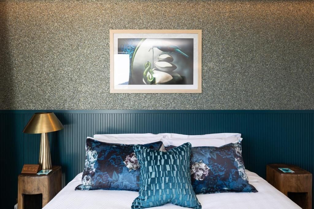 A neatly made bed with patterned pillows, a framed abstract painting above it, and a lamp on a wooden nightstand beside the bed.