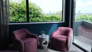 A modern room with two purple armchairs, a small side table with a plant, and a large window offering a view of greenery and buildings. - Tirohanga Nui (Oamaru)