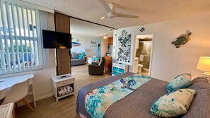 Quarto com cama, escrivaninha, televisão e vista para a sala de estar.