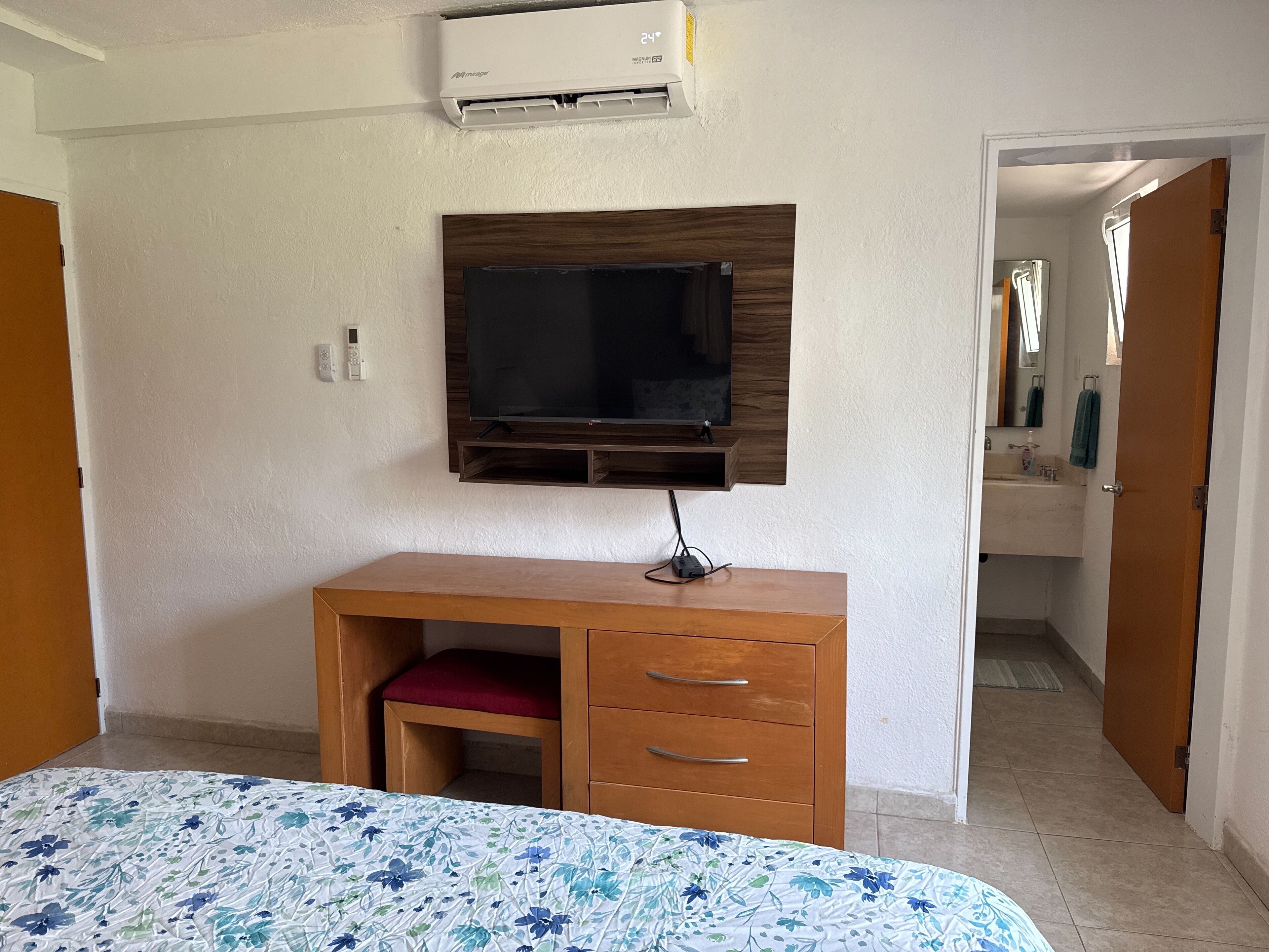 Ein Schlafzimmer mit einem Bett, einem hölzernen Schreibtisch und einem darüber montierten Fernseher. Durch eine offene Tür ist ein Badezimmer zu sehen.