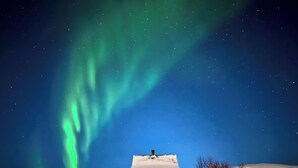A house with a snow-covered roof and a garage, surrounded by a snowy landscape under a night sky with visible stars and the Northern Lights.