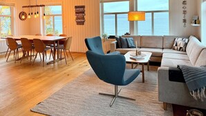 A spacious living area with a wooden dining table, blue chairs, a sofa, and a view of the sea through large windows.