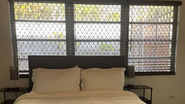 A bed with white linens, two bedside tables with lamps, and a window with a metal grid. - Quiet 1BR: 4 mins to Beach 10 mins to Downtown (Nassau)