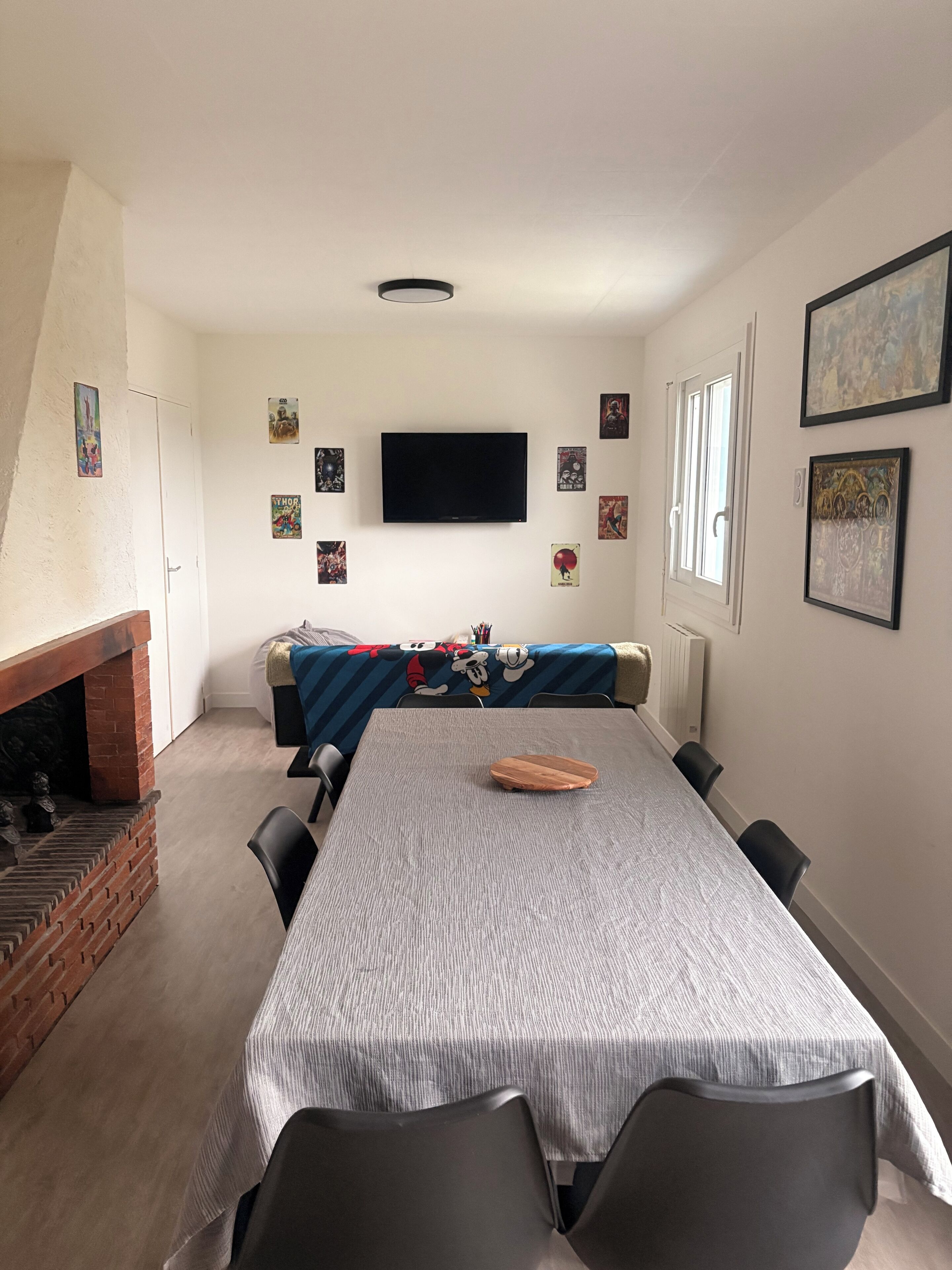 Een eethoek met een lange tafel, stoelen, een televisie en verschillende posters aan de muur.