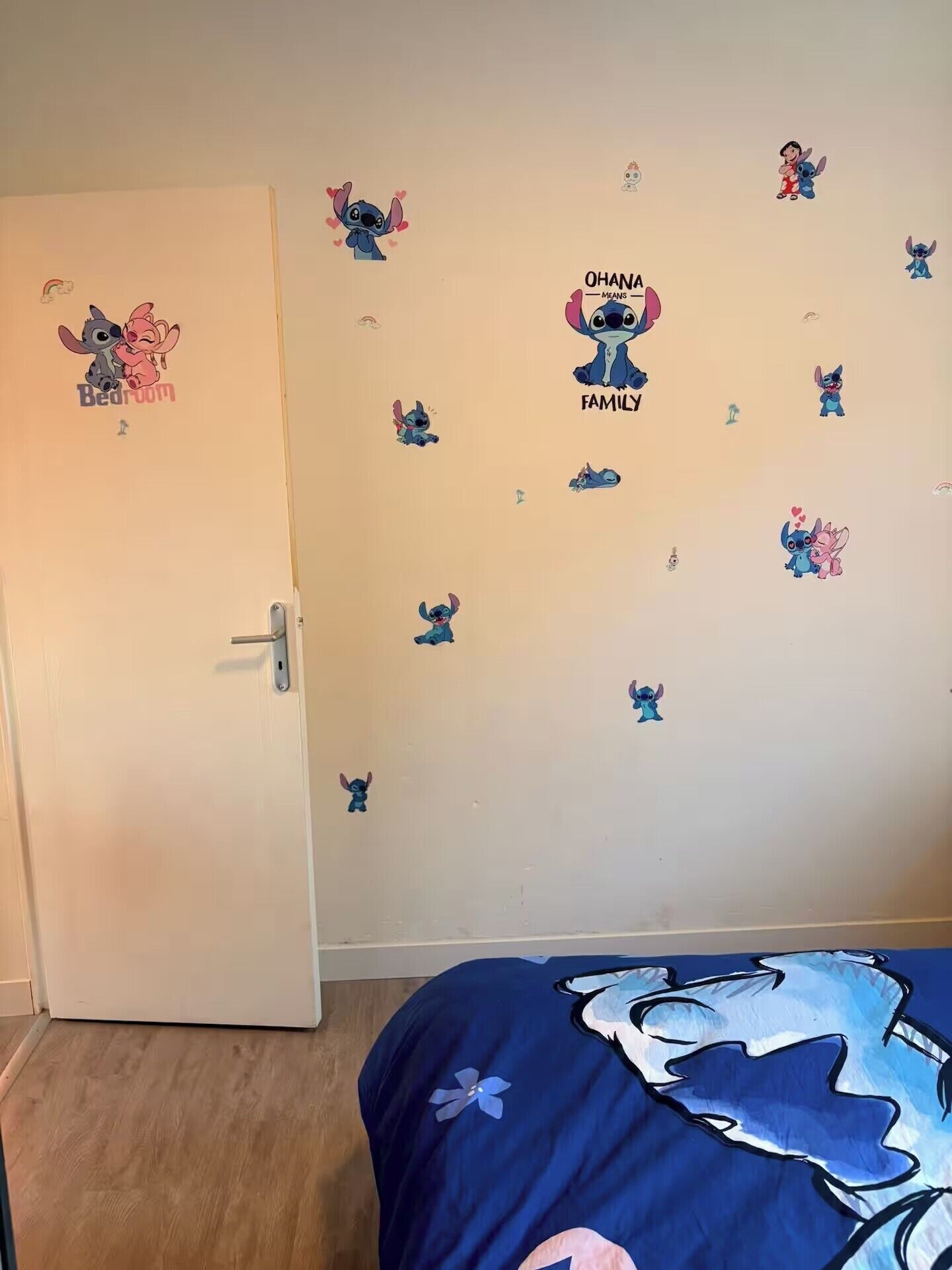 Une chambre avec une porte, un lit recouvert d’une couverture bleue et des murs décorés d’autocollants représentant Stitch et d’autres personnages.