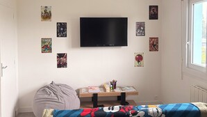 Una camera da letto moderna con un letto, un pouf, un tavolino con libri e articoli di cancelleria e una TV appesa al muro.
