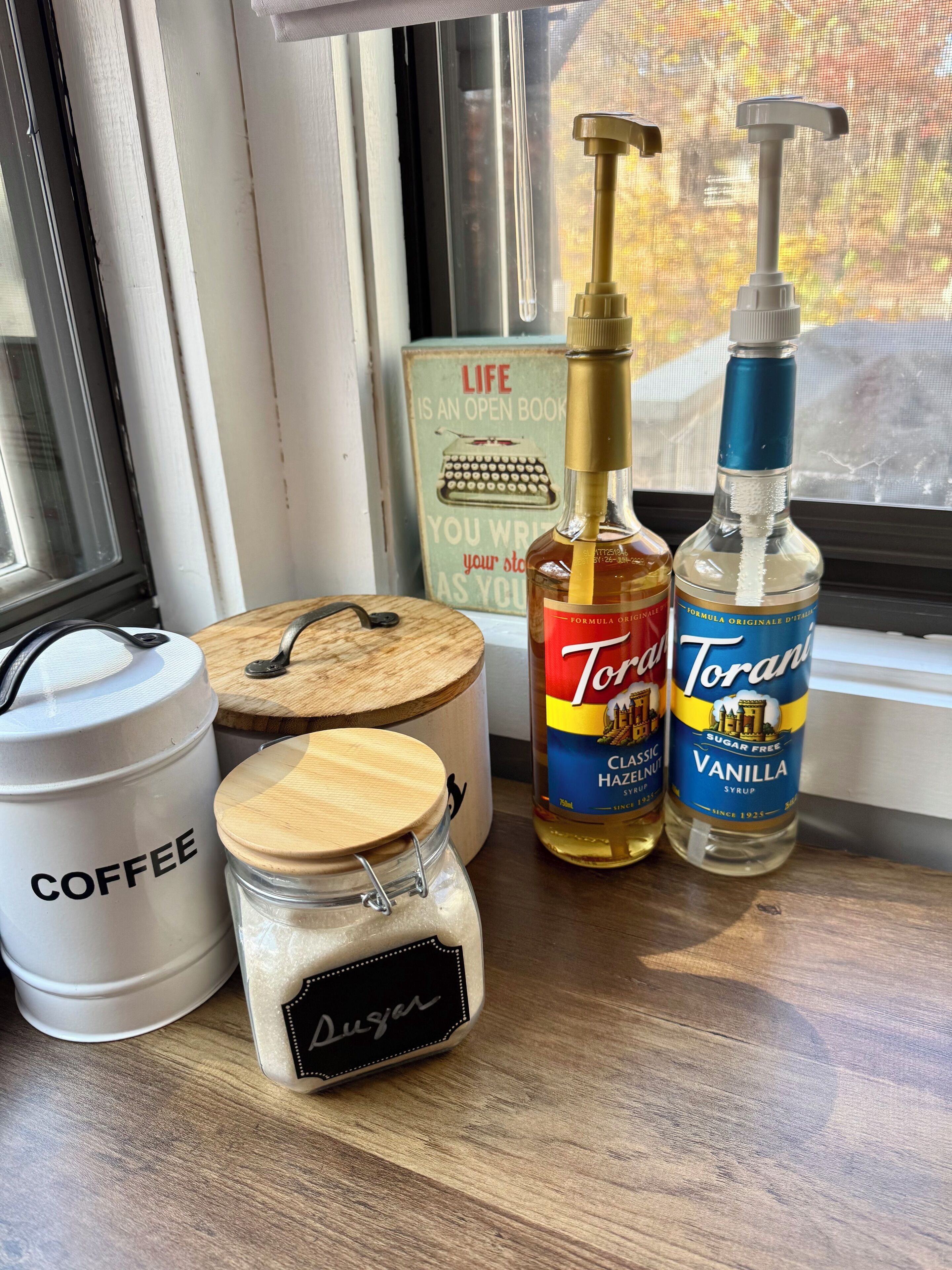 A window sill with a coffee container, a jar labeled 'Sugar', a Torani syrup bottle, and a vintage typewriter sign.