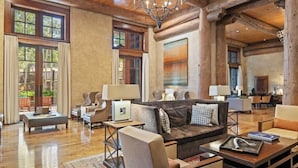 Ein geräumiges Wohnzimmer mit einer großen Couch, Sesseln und einem Couchtisch. Mehrere Fenster sorgen für natürliches Licht, und eine Kristallleuchter hängt von der Decke.