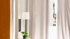 Una stanza con una porta in legno, una scrivania bianca con una lampada, una pianta e vista sugli edifici attraverso una finestra con tende.
