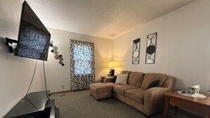 A living room with a sectional sofa, a flat-screen TV mounted on the wall, a lamp on a side table, and a window with patterned curtains. - Spacious 4-bedroom house in Fairmont (Fairmont)