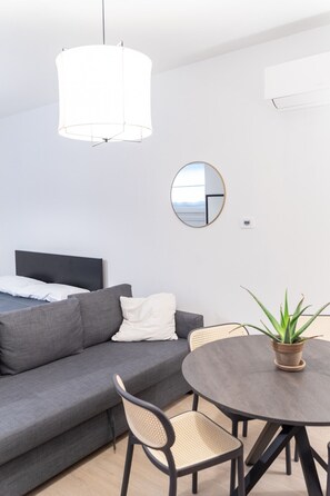 Una habitación moderna y minimalista con un sofá gris, un espejo redondo, una mesa de comedor con sillas y una lámpara colgante.