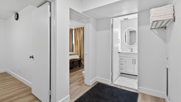 A modern hotel room with a wooden floor, a black rug, a bed with beige curtains, a bathroom with a mirror, and a towel rack. - 2BR | Warm | King Bed | Downtown | Expo Centre | Roger's Place (Edmonton)