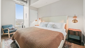 Una habitación de hotel moderna con una cama grande, vistas a la ciudad, una silla azul y una mesita.