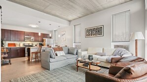 Una sala de estar moderna con un sofá gris, un sillón de cuero marrón y una mesa de centro. Se ve una cocina con armarios de madera oscura y un microondas al fondo.
