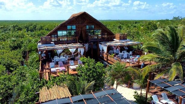 A wooden structure with multiple levels, including a balcony with white curtains and lounge chairs, surrounded by lush greenery and palm trees. - Casa Hayhu Beach (Mahahual)