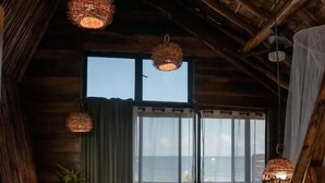 A rustic room with a high ceiling, woven light fixtures, a large window with a view, and a round woven table. - Casa Hayhu Beach (Mahahual)