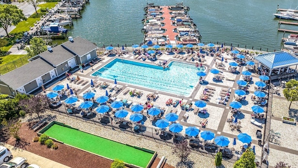 An aerial view of a resort with a large swimming pool surrounded by sun loungers and umbrellas, a dock with boats, and a river in the background. - Serenity in Ocean Pines | 1 Story Home with Backyard Grill (Ocean Pines)