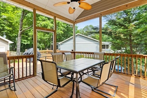 A covered outdoor deck with a table and chairs, surrounded by a wooden railing, overlooking a residential area with trees. - A Wave From it All | Screened Porch Outdoor Dining (Ocean Pines)