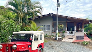 A red and white jeep parked in front of a house with a porch, a satellite dish, and a garden.