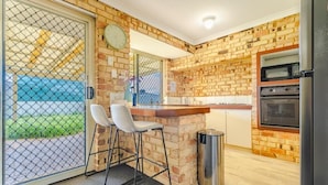 A kitchen with a brick wall, a microwave, an oven, a trash bin, and a seating area with two chairs. - StayAU 4BR Family Retreat with Garden (Seville Grove)